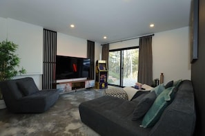 A modern living room with a flat-screen TV, a sofa, a gaming console, and a large window with curtains. - Chalet Le Select - Private Spa with River View (Les Éboulements)