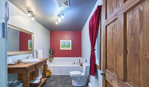 Un baño con tocador de madera, un lavabo blanco, un espejo, una pared roja, un cuadro, una bañera y una puerta de madera.