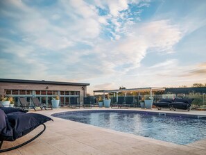 Une piscine avec des chaises longues, un espace de détente couvert et un bâtiment doté de grandes fenêtres.