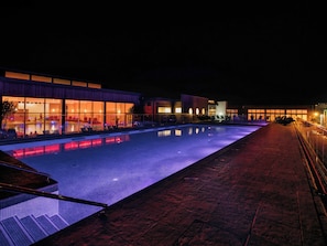 Ein Poolbereich bei Nacht mit Glasveranda, überdachten Sitzgelegenheiten und einem Gebäude mit beleuchteten Fenstern.