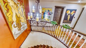 Una escalera curva con un candelabro, un cuadro grande y un cartel en la pared.