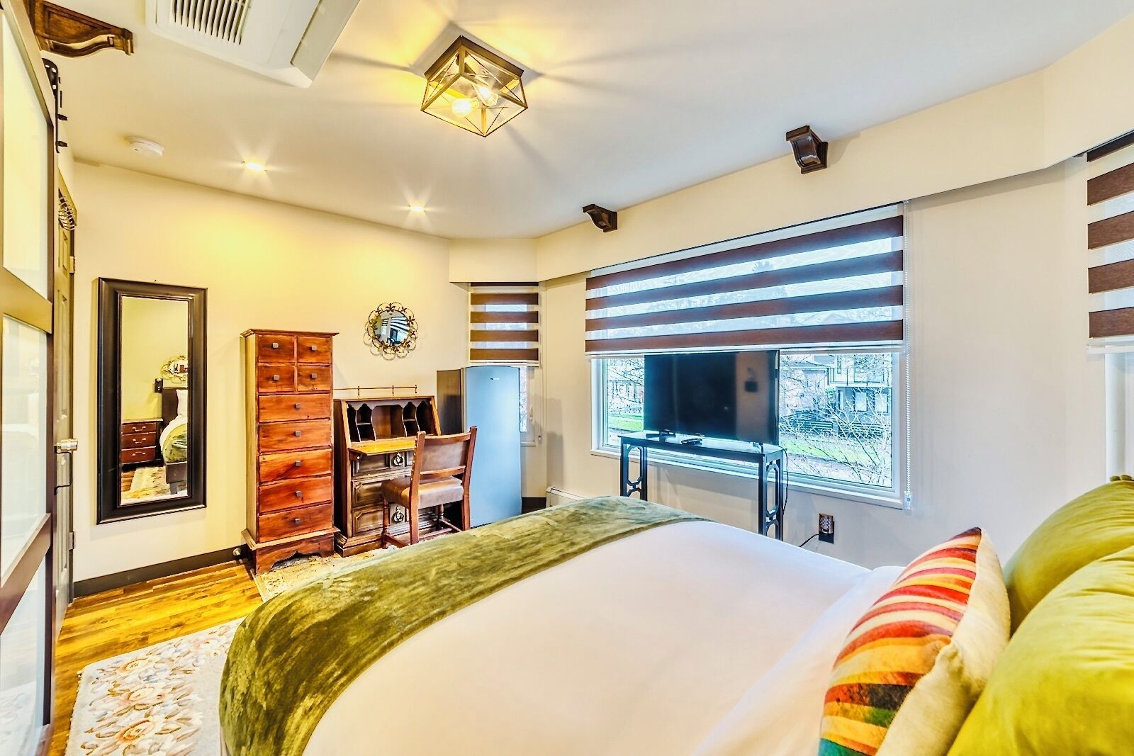 Ein Schlafzimmer mit Bett, Kommode, Spiegel, Kleiderschrank, Fernseher und einem Fenster mit Jalousien.