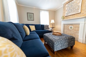 A living room with a blue sofa, patterned ottoman, and a fireplace with a brick wall.