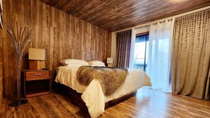 Une chambre avec un plafond en bois, un lit recouvert d’un plaid en fourrure, une table de chevet et une lampe.