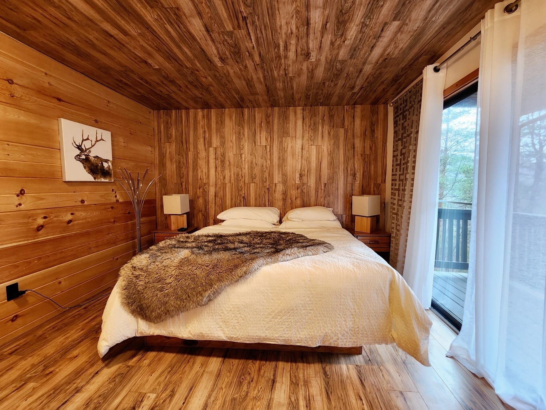 A wooden cabin bedroom with a bed, two bedside lamps, a deer head artwork, and a window with curtains.