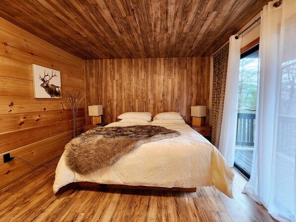A wooden cabin bedroom with a bed, two bedside lamps, a deer head artwork, and a window with curtains.