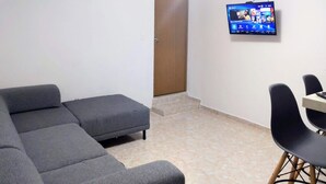 A room with a grey sectional sofa, a small dining table with black chairs, a flat-screen TV mounted on the wall, and a door leading to another room.
