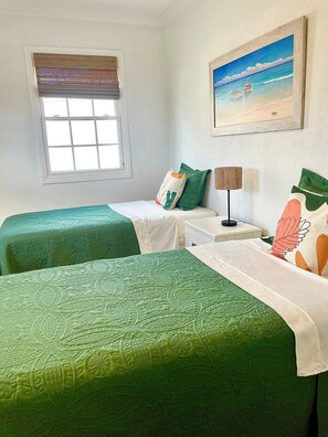Ein Zimmer mit zwei Betten, einem Fenster mit Jalousien, einer Lampe, einem Strandbild und einem Nachttisch.