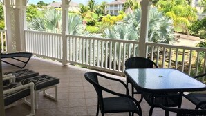 A covered outdoor area with a table and chairs, a lounge chair, and a view of a residential neighborhood with houses and palm trees.
