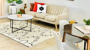 A modern living room with a beige sofa, a round coffee table, and a dining table with chairs. There are three wall-mounted decorative panels above the sofa.
