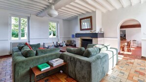 Ein geräumiges Wohnzimmer mit einer grünen Sofagarnitur, einem Kamin und Blick ins Freie durch große Fenster.