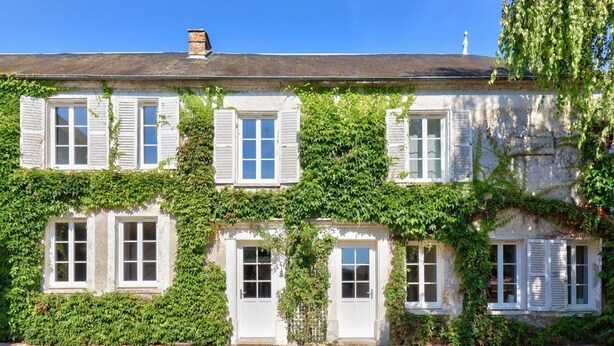 Ein zweistöckiges Haus mit weißen Fensterläden und einer Stein-Fassade, teilweise bewachsen mit Efeu.