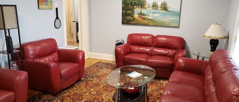 A living room with red leather sofas, a glass coffee table, a patterned rug, and a painting of a tropical beach scene.