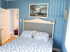 Une chambre à coucher avec un lit, un tableau représentant un paysage marin et une porte donnant sur une autre pièce.
