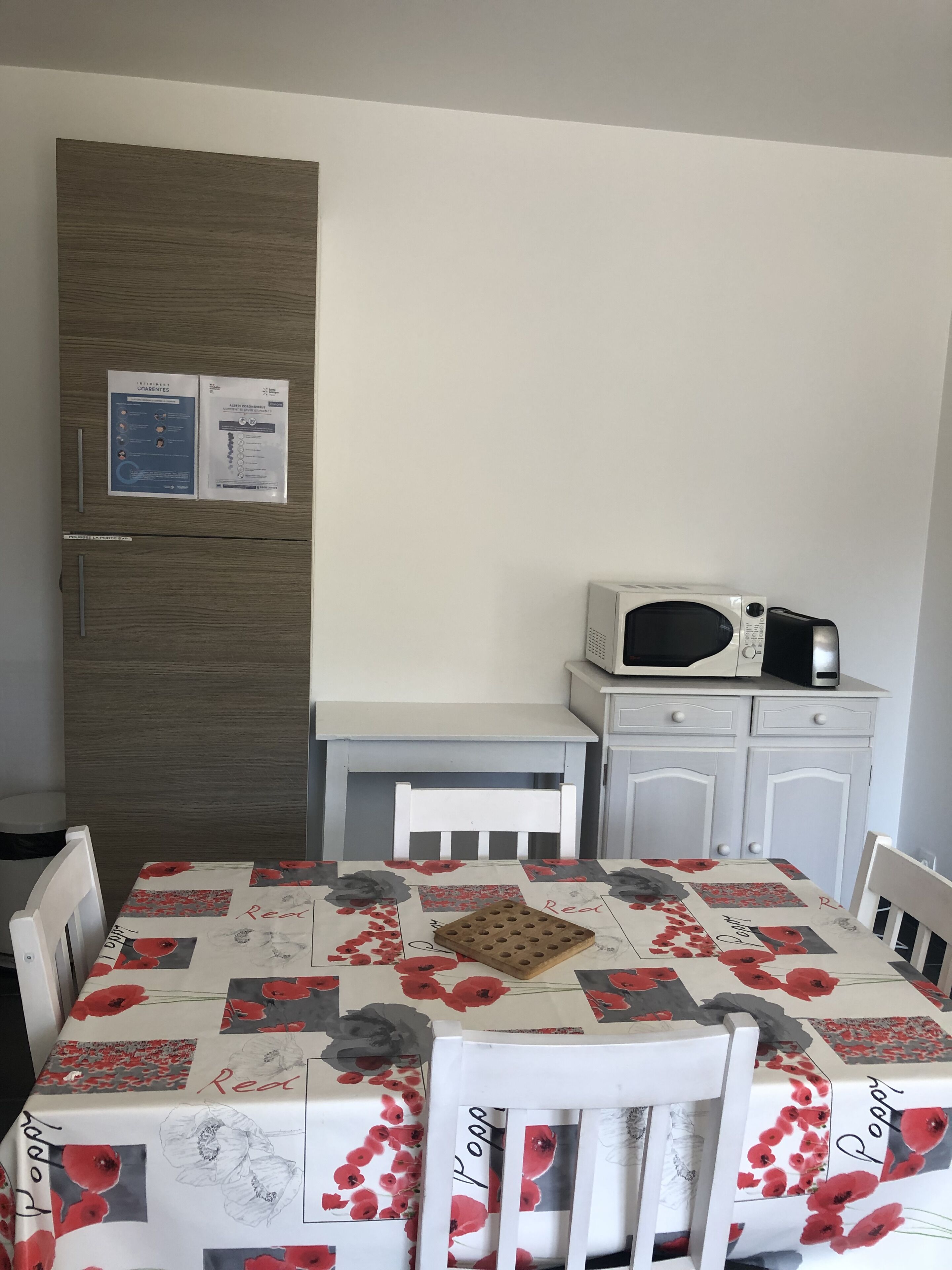 A dining area with a table covered in a floral tablecloth, a white chair, a microwave on a cabinet, and a wooden door with papers on it.