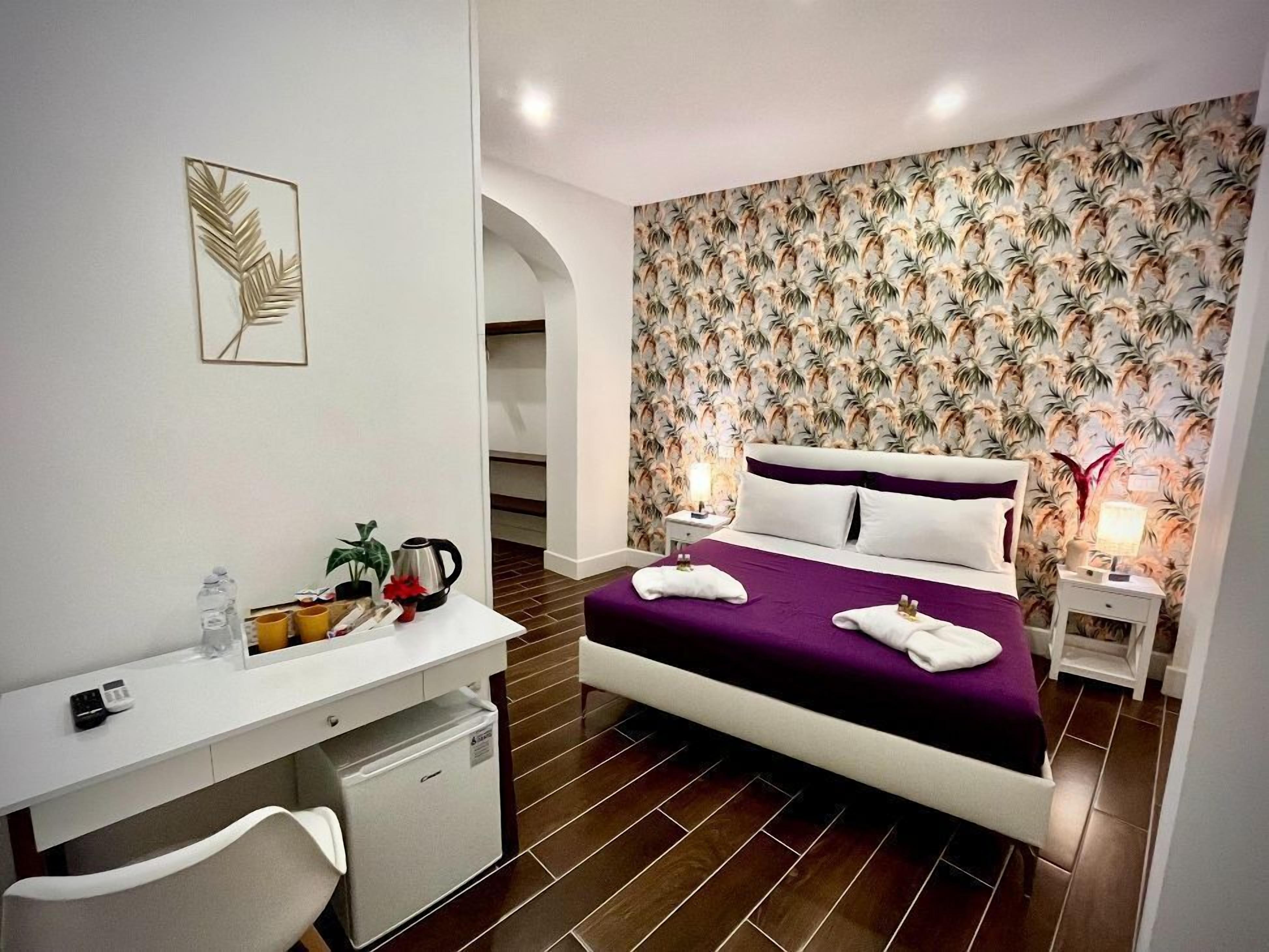 A modern hotel room with a bed, a desk, a chair, a small fridge, a nightstand, and a wall with a tropical leaf pattern.