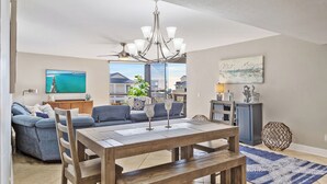 A modern living room with a wooden dining table, blue sofas, a chandelier, and a large window offering a view of the city. - Mainsail 332 - Updated Beachfront Condo, Free Bch Svc, Pool, Hot Tub, Tennis! (Miramar Beach)