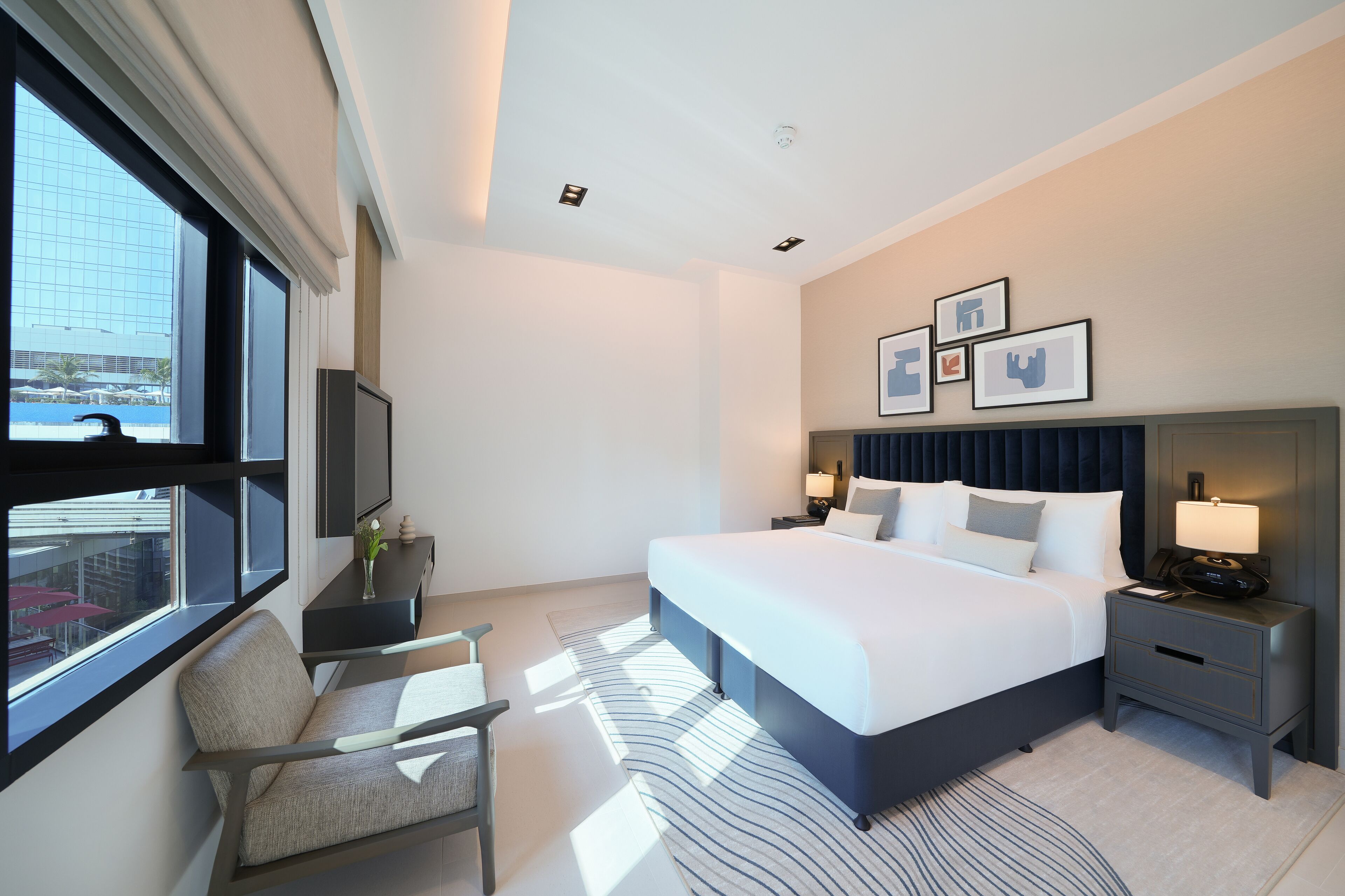 Quarto de hotel moderno com uma cama grande, mesas de cabeceira, uma cadeira e vista da cidade por grandes janelas.