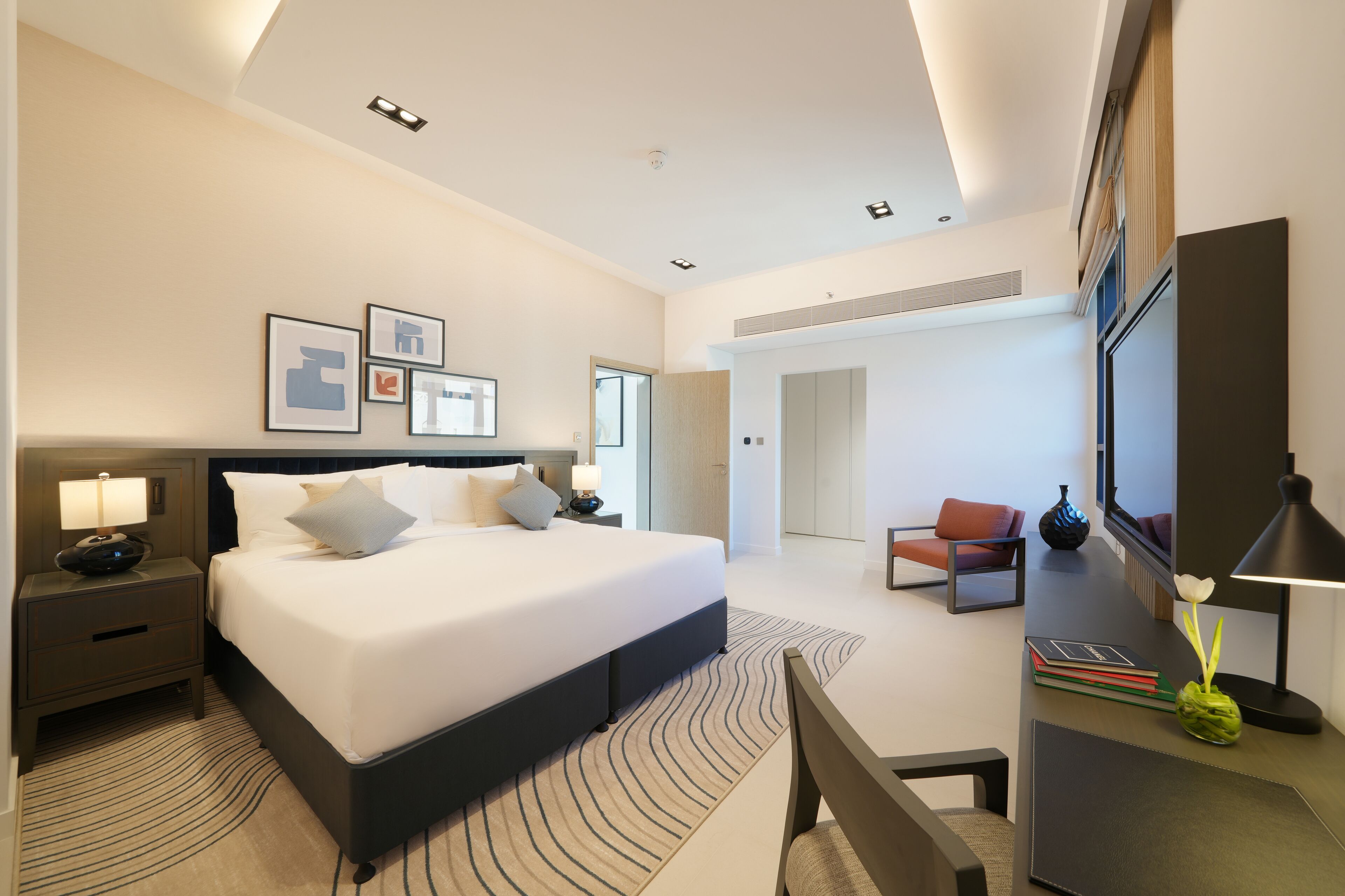A modern hotel room with a large bed, bedside tables, a desk, and a chair. There is a television and a vase with flowers on the desk.
