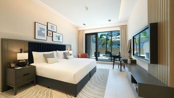 A modern hotel room with a large bed, a bedside table with a lamp, a flat-screen TV mounted on the wall, and a sliding glass door leading to a balcony with a view of palm trees and buildings.