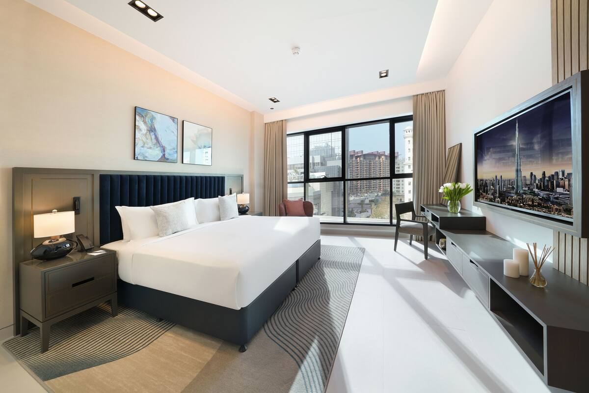 Une chambre d’hôtel moderne dotée d’un grand lit, de tables de chevet, d’un bureau, d’une chaise, d’une grande fenêtre donnant sur la ville et d’œuvres d’art accrochées aux murs.