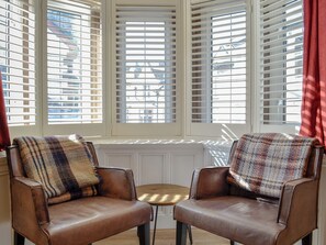A room with two leather armchairs facing a bay window with white shutters, a small wooden table, and a view of buildings outside.