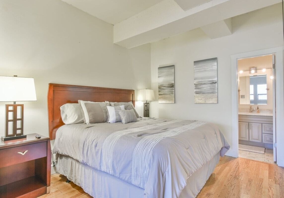 A bedroom with a large bed, a nightstand with a lamp, and a bathroom visible through an open door.