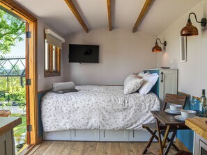 Quarto compacto com cama, televisão, uma mesinha com tigelas e uma taça de vinho, e vista pela janela.