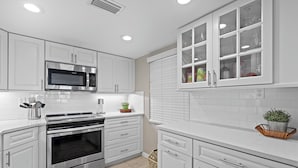 A modern kitchen with white cabinetry, stainless steel appliances, and a window with blinds. - 🌴Ocean View Condo on No-Drive Beach-Castle Reef 312🌴 (New Smyrna Beach)