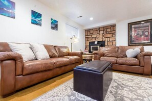 Ein Wohnzimmer mit zwei braunen Ledersofas, einem schwarzen Hocker und einem gemusterten Teppich. An den Wänden hängen gerahmte Bilder und ein Kamin aus Stein.