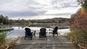 A wooden deck with three Adirondack chairs and a small table overlooking a lake, surrounded by autumn foliage and a cloudy sky. - Babcock Lake Luxury Retreat: Direct Waterfront, Deck & Huge Game Room (Petersburgh)