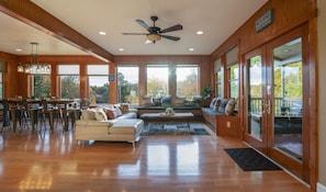 A spacious living room with a large sofa, a dining area with bar stools, and a view of a lake through large windows. - Babcock Lake Luxury Retreat: Direct Waterfront, Deck & Huge Game Room (Petersburgh)