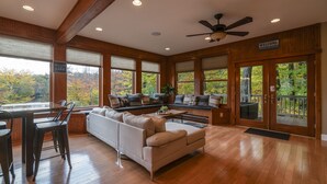 A spacious living room with a large sofa, a dining area with chairs, and a view of autumn trees through large windows. - Babcock Lake Luxury Retreat: Direct Waterfront, Deck & Huge Game Room (Petersburgh)