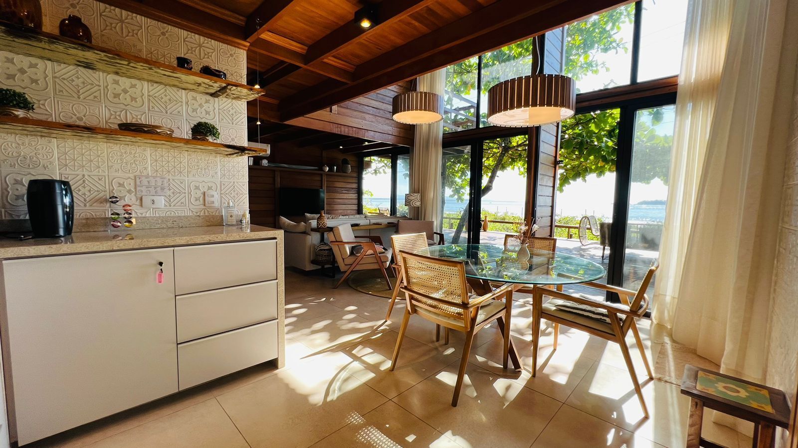 A modern kitchen with a glass dining table, wooden chairs, and a view of the sea.
