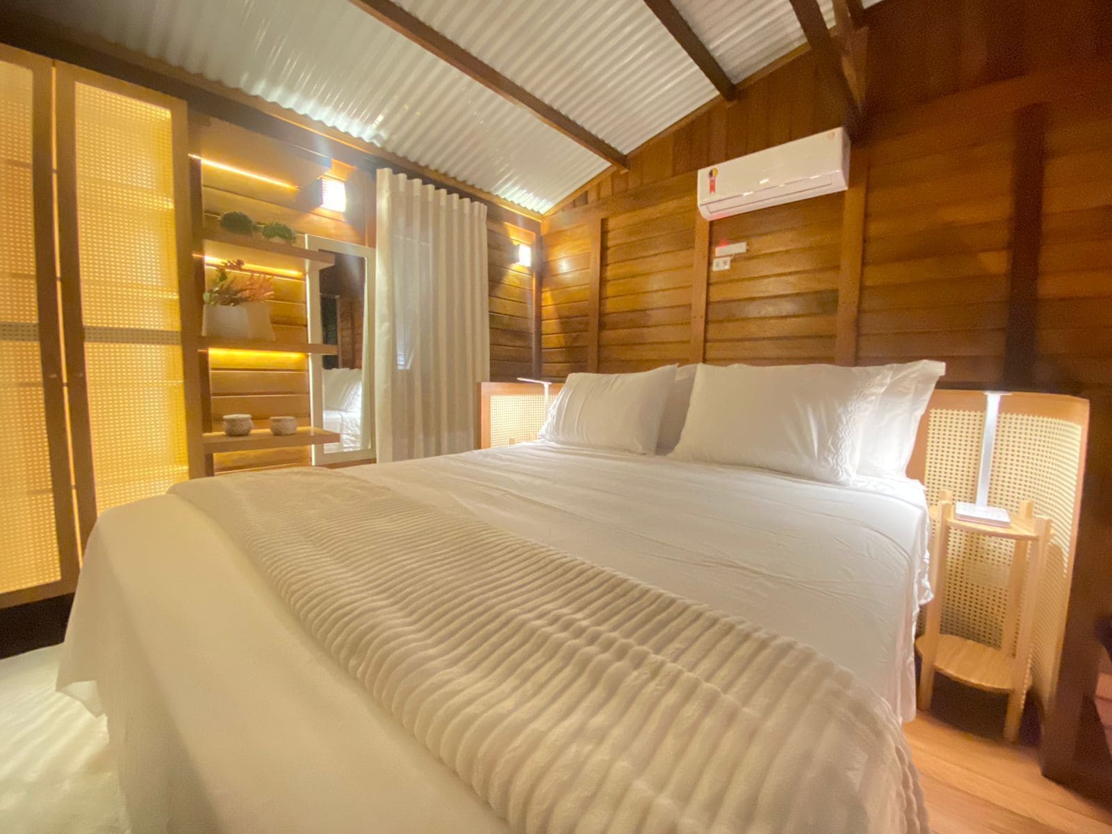 Quarto tipo cabana de madeira com uma cama grande, uma mesa de cabeceira, uma luminária e uma janela com cortinas.