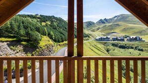 A wooden balcony with a view of a winding road, lush green hills, and a cluster of buildings in the distance. - Floor -1, view ski slopes, jacuzzi, hammam, balcony, parking, tv, 30m², Peyragudes (Germ)
