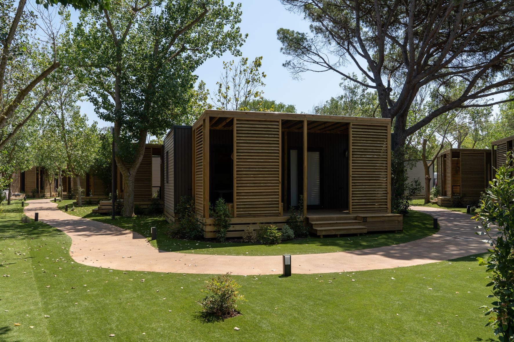 A row of wooden cabins with a paved walkway between them, surrounded by trees and grass.