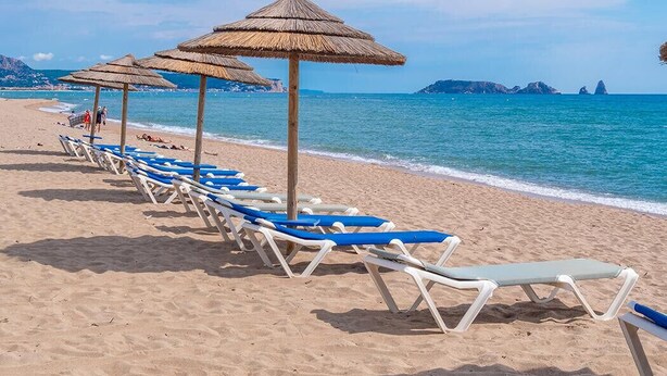 Una playa con filas de tumbonas y sombrillas de paja, un mar azul cristalino y formaciones rocosas a lo lejos.