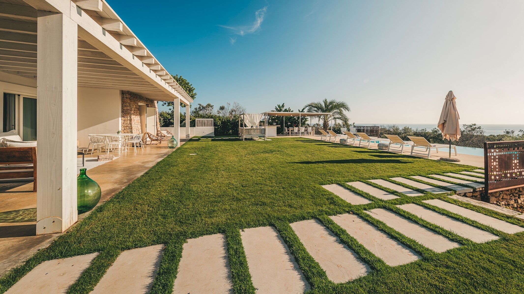 A well-maintained lawn with a stone pathway leading to a covered seating area, overlooking a pool and ocean view.