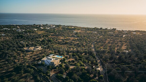 An aerial view of a coastal area with scattered buildings, a prominent white building with a blue roof, and a road leading towards the sea.