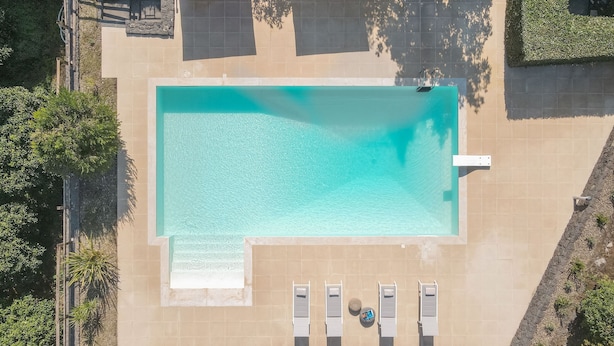 An aerial view of a rectangular swimming pool with a tiled deck surrounding it. There are lounge chairs aligned along the pool's edge and a small round table with a bottle and a cup. - Villa Kimia 8, Emma Villas (Acireale)