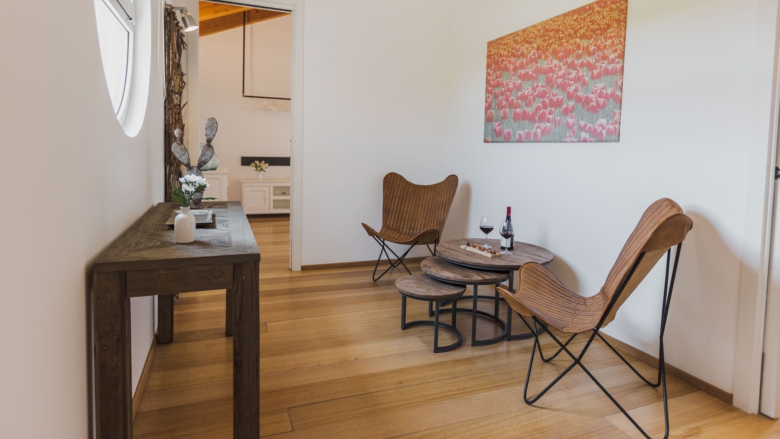 A dining area with a wooden table and two chairs, a small round table with a bottle of wine, and a painting on the wall.