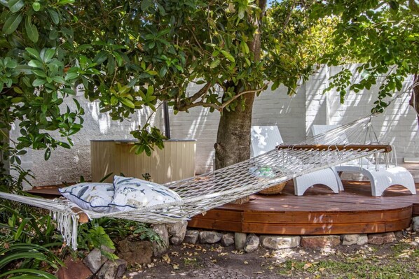 A hammock on a wooden deck with a white chair, surrounded by greenery and a white wall.