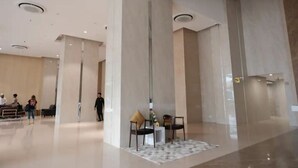 Eine geräumige Lobby mit hohen Decken, großen Säulen und einem polierten Boden. Im Hintergrund sind einige Personen zu sehen, und es gibt eine kleine Sitzecke mit einem Teppich.