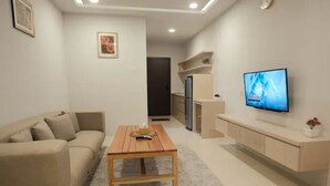 Una sala de estar moderna con un sofá color beige, una mesa de centro de madera, un televisor sobre un mueble blanco y una cocina al fondo.