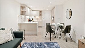 Una cocina moderna con armarios blancos, un comedor con mesa y sillas de vidrio, y una sala de estar con sofá y alfombra azul.