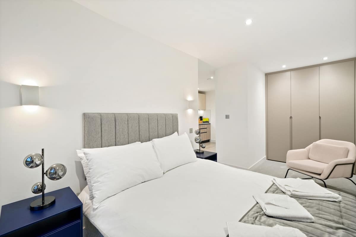A modern hotel room with a large bed, a bedside table with a lamp, a chair, and a wardrobe.