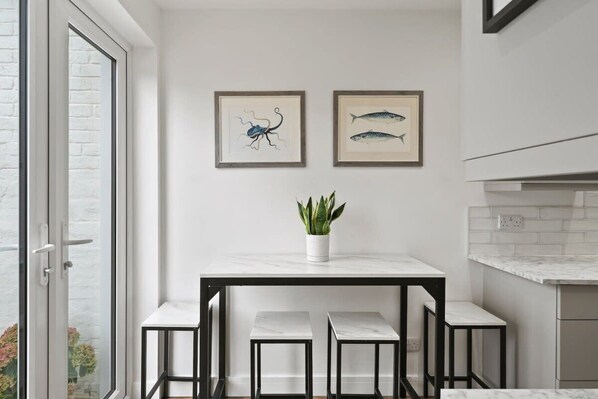 Une cuisine moderne avec un plan de travail en marbre blanc, des tabourets en métal noir et deux illustrations encadrées représentant des poissons, accrochées au mur.