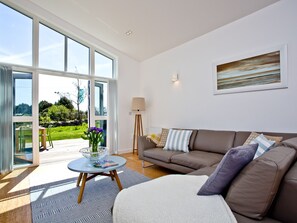 A modern living room with a brown sofa, a round coffee table, and a large window offering a view of a garden. - 2 bedroom accommodation in Carbis Bay, near St Ives (Carbis Bay, near St Ives)