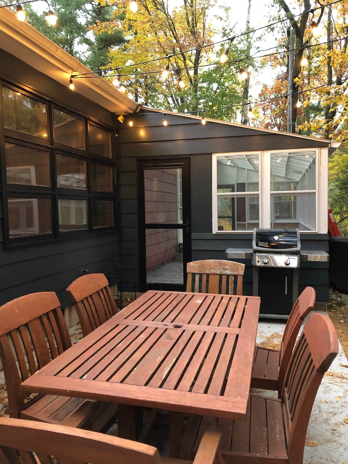 Une terrasse avec une table et des chaises en bois, un barbecue et des guirlandes lumineuses.